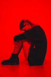 A figure sits curled with their arms wrapped around their knees against a vivid red backdrop, showcasing a contemplative and introspective moment.